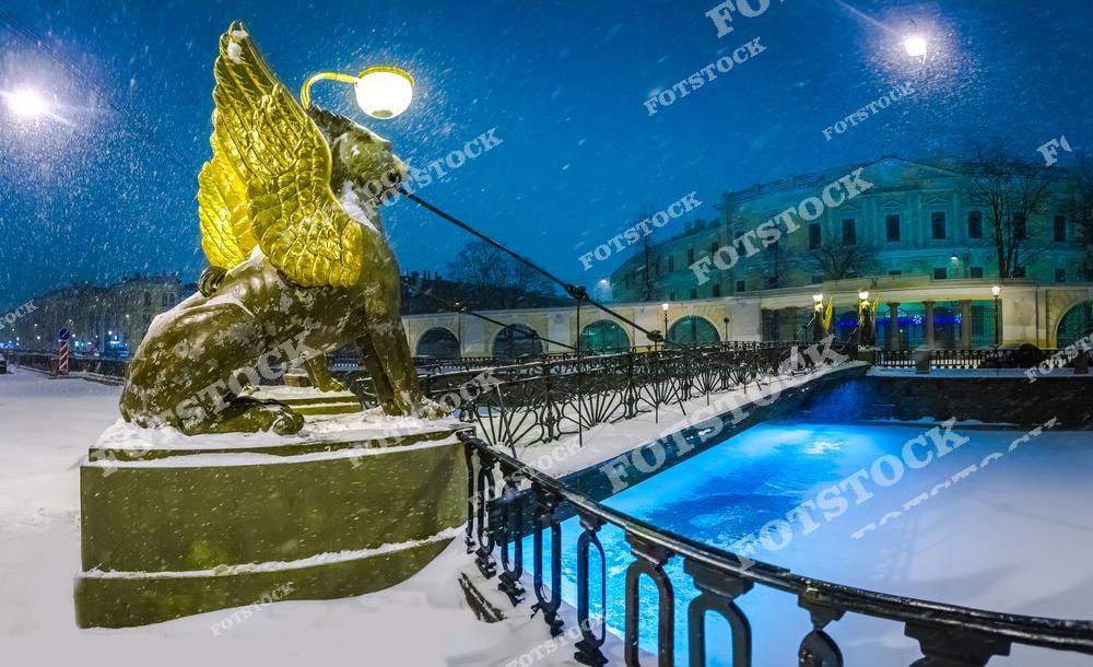 На фотографии изображён Банковский мост через канал Грибоедова в зимнюю ночь. Золотой грифон с крыльями, освещённый фонарями, подчёркивает атмосферу мистики и красоты. Снегопад и замёрзший канал придают сцене ощущение зимней сказки. Этот мост — одна из самых узнаваемых достопримечательностей Санкт-Петербурга, известная своей изысканной архитектурой.